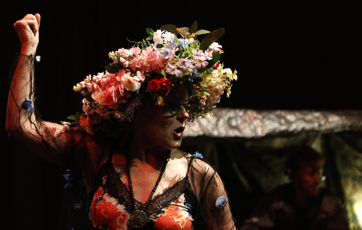 An older woman wearing a headdress of flowers, floral slip dress, and medallion, holding her fist aloft.