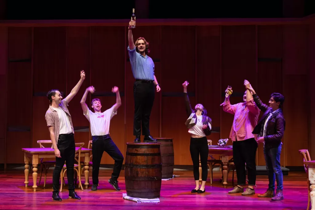 A man stands on a barrel raising a bottle to the air, surrounded by other characters from the show.